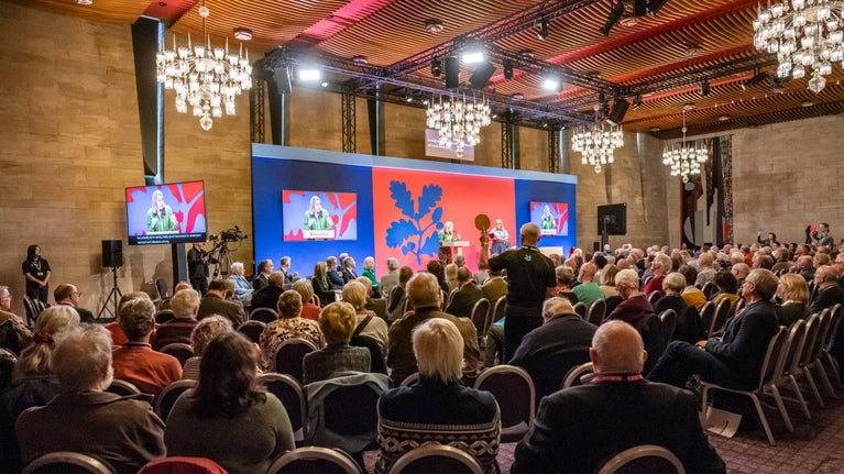A seated audience watch a lady speaking at a podium in front of a brightly coloured backdrop with chandeliers overhead